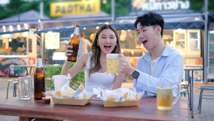 Young couple enjoying drinks and fried snacks at an outdoor street food market, laughing and raising their glasses.