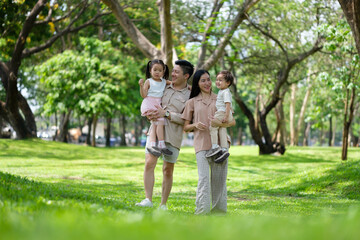 Fototapeta premium Happy family walking together in a sunny green park, each parent carrying a child.