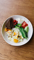 Top view of healthy breakfast served on white ceramic plate with fried eggs, fresh vegetables including tomatoes and greens, and slices of dark bread.