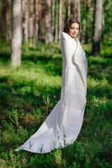 A young woman stands in a summer forest wrapped in a warm blanket. Girl wakes up in a summer forest. Eco-comfort and relaxation © Oleg Gekman