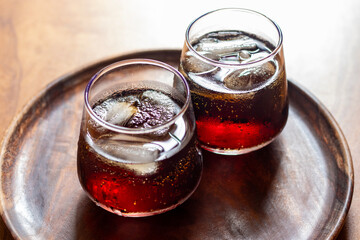 Two glasses of dark soda with ice cubes sit on a wooden serving board, placed on a wooden table under warm lighting.
