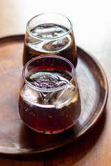 Two glasses of dark soda with ice cubes sit on a wooden serving board, placed on a wooden table under warm lighting.
