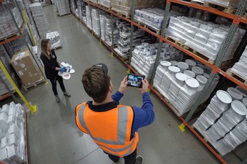 Warehouse inventory with digital technology. Two people use technology to track stock. Shelves filled with various packaged items