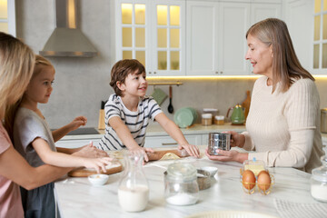 Little kids with their mother and grandmother making dough at white marble table in kitchen