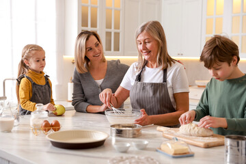Little kids with their mother and grandmother making dough at white marble table in kitchen