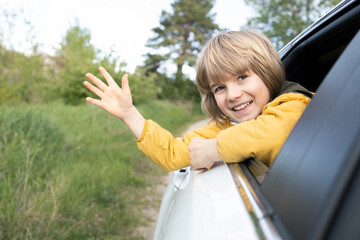 A happy boy looks out of the car window and happily greets or says goodbye to someone. Hello travel, adventure, vacation. On vacation by car. Mood for an interesting pastime