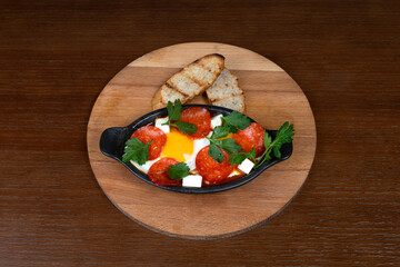 Shakshuka with pepperoni and feta cheese with greens, egg and rye bread on the table in the restaurant on a wooden stand