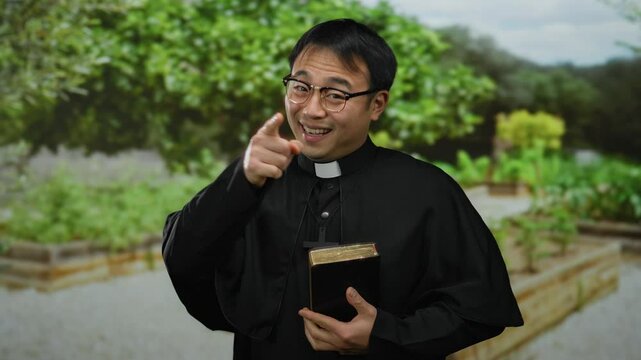 Asian priest smiling in outdoor park holding bible with cross necklace, trees in background, wearing black clergy attire, glasses, daylight setting, spiritual, peaceful atmosphere.