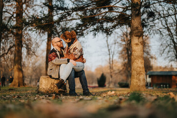 A couple sits together on a tree stump in a serene park with autumn colors, expressing emotional intimacy. Trees and dried leaves surround the background, creating a peaceful natural setting.