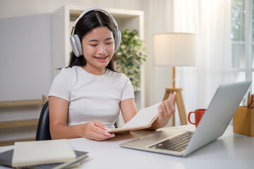 Asian teenage girl smiling while taking notes and studying online with a laptop at home.