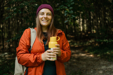 Woman in hiking gear holding a yellow thermos bottle and smiling in a forest. Represents...