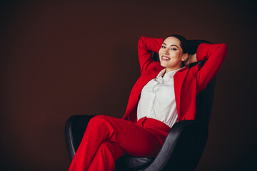 Confident Businesswoman in Red Suit Relaxing in Office Chair on Brown Background