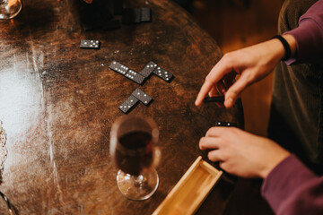 Close-up of hands playing dominoes on a wooden table, accompanied by a glass of wine. The setting...