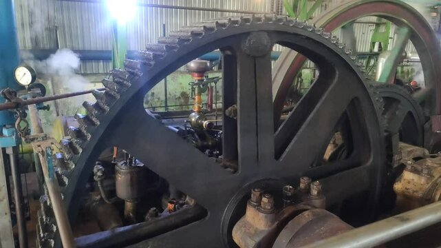 Large flywheel rotates in an old steam-powered factory engine room with visible gears, pipes, and steam.