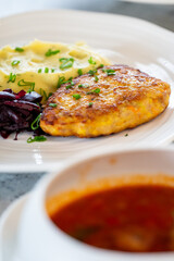 A close-up of a plated meal featuring mashed potatoes with herbs, breaded fried meat, sliced beets, and a bowl of tomato-based soup, presented on a rustic wooden table.