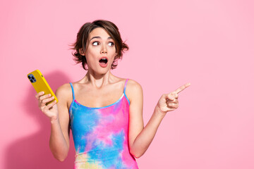Charming woman with brunette hair points in excitement while holding a yellow smartphone, summer...