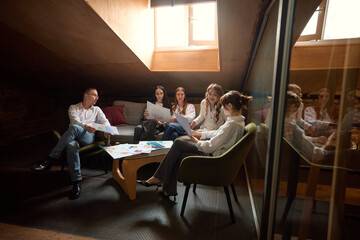 Business team reviews documents in office lounge area, sitting on sofa and chairs, discussing strategy. Marketing review. Concept of teamwork, analysis, strategy, meeting, collaboration.