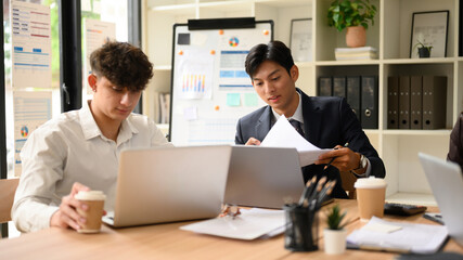 Two business professionals analyzing documents and working on laptops during a meeting in a modern office. Focused teamwork with charts and coffee cups on the table