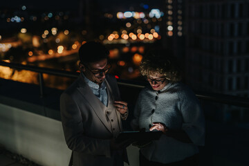 Fototapeta premium Two colleagues brainstorm over a tablet on a city rooftop at night, amidst glowing city lights, showcasing a collaborative and focused work environment with a modern urban backdrop.