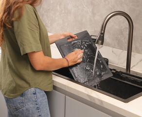 Woman washing a black cutting board under running water in a modern kitchen sink. Hygienic cleaning, kitchen routine, and household chores concept.
