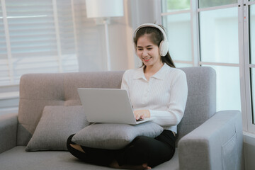 Naklejka premium Asian woman living lifestyle wearing wireless headphones staring at laptop screen and communicating with clients freely on sofa at home.