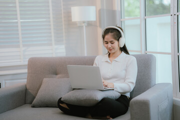 Naklejka premium Asian woman living lifestyle wearing wireless headphones staring at laptop screen and communicating with clients freely on sofa at home.