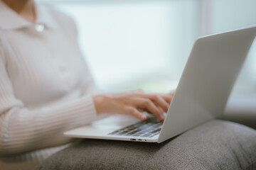 Asian woman living lifestyle wearing wireless headphones staring at laptop screen and communicating with clients freely on sofa at home.