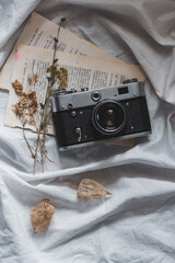 An old film camera lies on an old book on a white crumpled sheet, around dry leaves of plants, herbarium flowers. Flat lay.