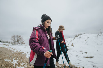 Two women hike in a wintry countryside, dressed warmly with trekking poles, enjoying the nature and...