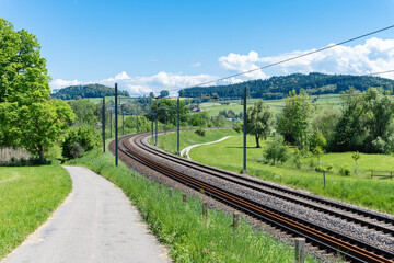A winding railway line through a green landscape on a sunny day, surrounded by trees and hills.