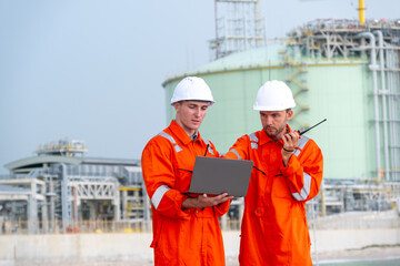 Two workers in safety gear collaborate on a laptop at an industrial site near a large storage facility