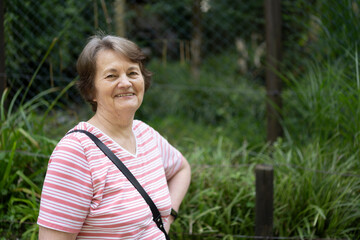 An elderly gray-haired woman in a striped T-shirt stands smiling against the background of a green...