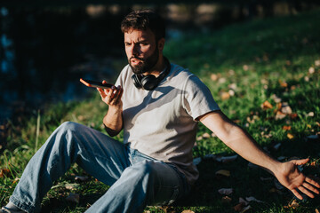 A young man with headphones uses a smart phone voice assistant while sitting in a grassy outdoor...