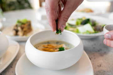 A close-up of a hand sprinkling fresh herbs into a bowl of soup, with other dishes blurred in the background, creating a cozy and appetizing dining scene