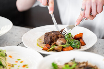 A person cuts a piece of cooked meat on a white plate, accompanied by mushrooms, carrots, and greens. The gourmet dish is elegantly presented in a fine dining setting.