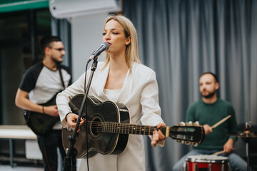 A female guitarist and singer performing alongside a band in a music studio setting. The rehearsal...