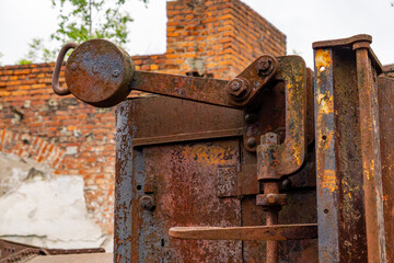 Close-up of an old rusty industrial lever with visible corrosion and weathering. In the background, there are brick ruins, emphasizing an abandoned and historical industrial setting.