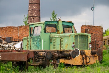 An old, rusty green-yellow shunting locomotive standing on overgrown railway tracks amid brick...