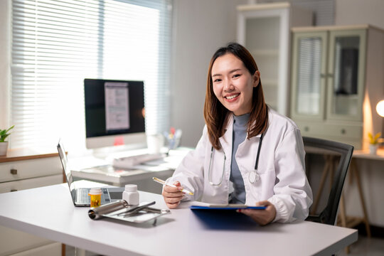 A woman in a white lab coat is sitting at a desk with a clipboard and a laptop - Powered by Adobe