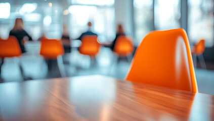 Close-up of an orange chair and wooden table in a blurred office environment with people sitting at desks near a window, creating a modern workspace atmosphere