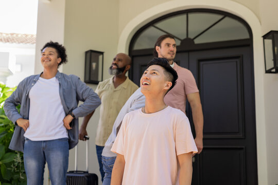 Looking diverse male friends standing on porch of home, with rolling suitcase and lantern sconces