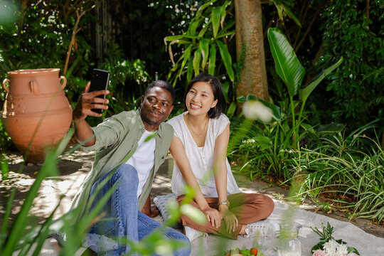Taking selfie diverse couple sitting on picnic blanket on patio, with smartphone and fruit bowl - Powered by Adobe