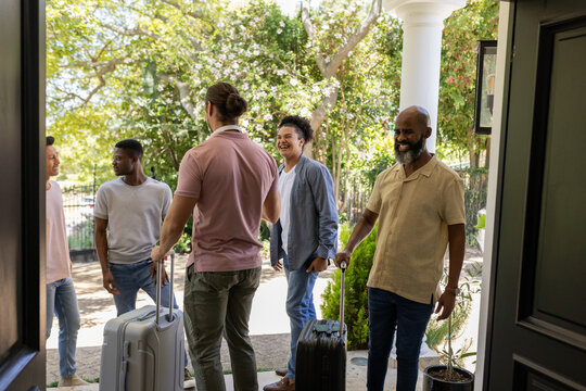 Arriving diverse male friends chatting on front porch, with rolling suitcases and potted plants - Powered by Adobe