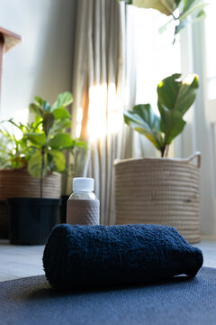 Rolled navy towel resting on dark exercise mat with water bottle, leafy plants near beige curtains