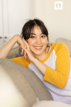 Leaning Asian woman resting chin on hands on beige living room couch, with mustard raglan shirt
