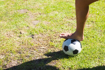 bare foot pressing soccer ball on patchy grass field under bright sunlight, casting sharp shadow