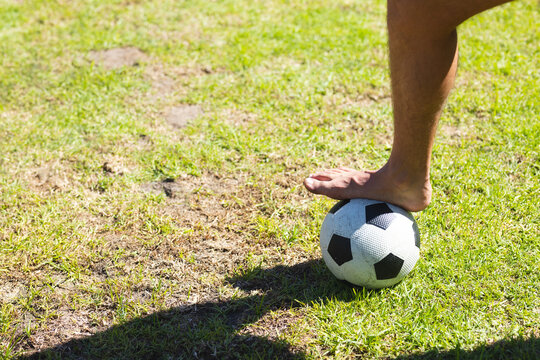 bare foot pressing soccer ball on patchy grass field under bright sunlight, casting sharp shadow