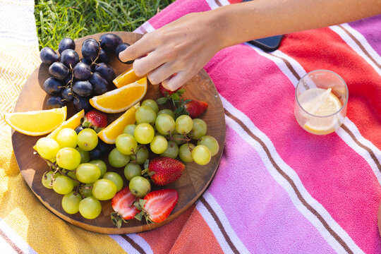 Reaching Asian woman picking orange slice on picnic blanket at park, with fruit board and juice