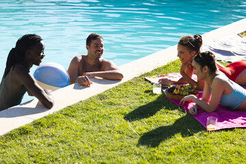 Chatting diverse friends lounging poolside on pink towel, with beach ball and fruit platter