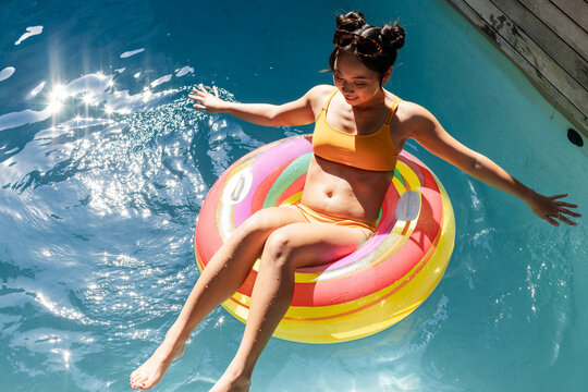 Floating Asian woman wearing yellow bikini on inflatable ring in backyard pool, with sunlit water